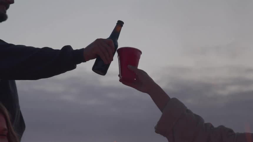 A hand with a plastic cup and a hand with a beer bottle make a toast outdoors as smoke rises in front of them