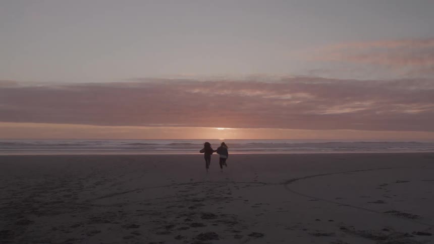 Two individuals holding hands run along a sandy beach towards the ocean during a sunset the sky is a warm soft blend of pink and orange casting a serene glow over the water and sand