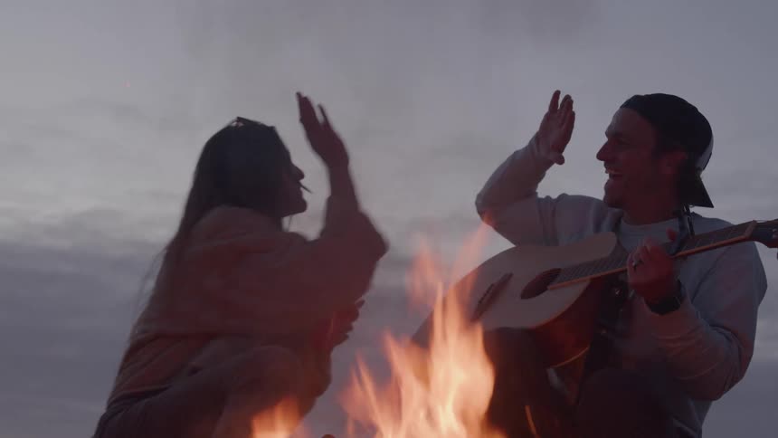 A couple sits by a campfire as the man plays a guitar creating excitement while the young woman laughs and holds a red cup on a cloudy evening