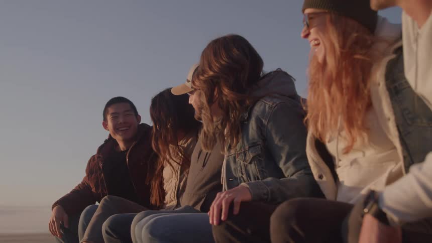 Small group of friends consisting of young adult males and females sitting and laughing at the beach outdoors during dusk or dawn