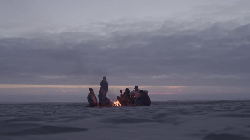 Group of friends sitting on the sand around a bonfire on a beach at dusk
