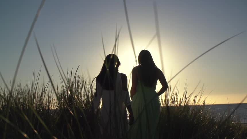 Two women in white dresses walking through tall grass next to a body of water as the sun sets