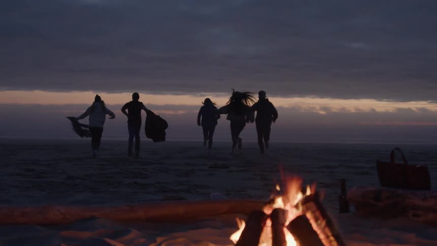 Group of friends running towards the sea on the beach at dusk