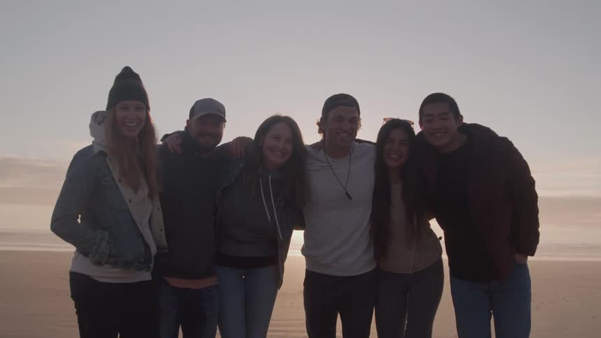 Group of six friends hugging on the beach at sunset