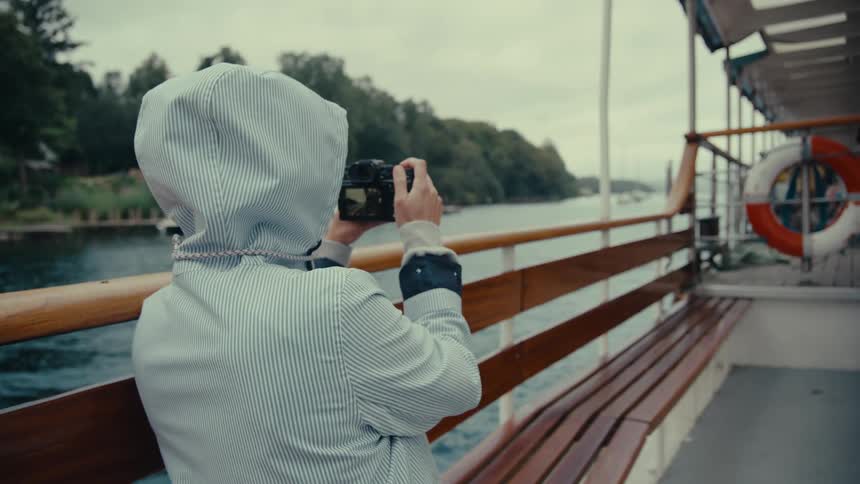 A person wearing a striped jacket with a hood is seen from behind holding a camera up to their face to take a photo they are on the deck of a boat with a large body of water and a lush treelined coast in the background