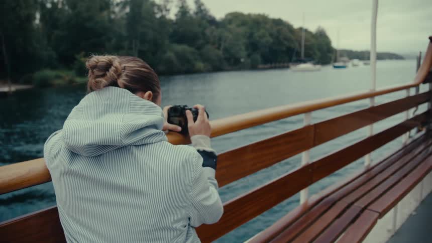 A young woman stands on the deck of a boat looking out at the water and distant treecovered hills she holds a camera and rests her arms on the wooden railing as she takes a photo of the view