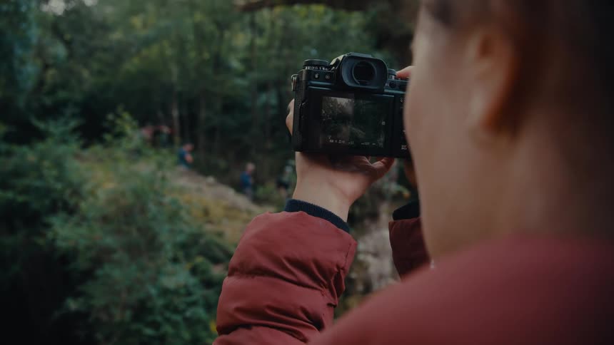A woman in a rust colored puffy jacket stands on a trail and holds a digital camera with a screen to take a photograph of some people on a rocky path below with a wooded area in the distance