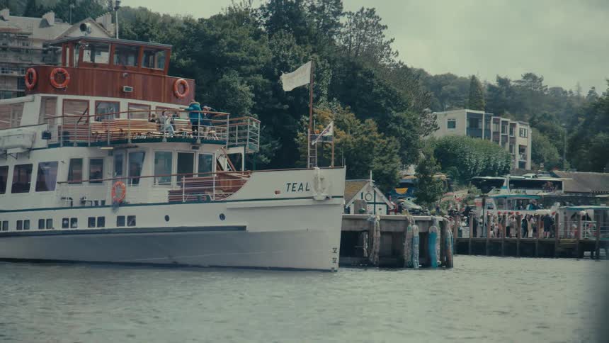 A boat with the name teal on its side floats on water next to a pier with people and trees in the background