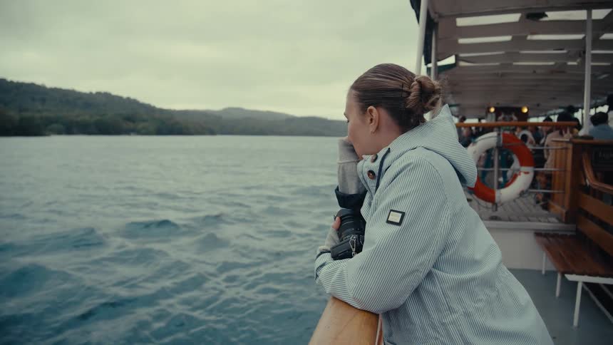A young woman stands on the deck of a boat looking out at the water and distant treecovered hills she holds a camera and rests her arms on the wooden railing as the boat moves across the choppy water