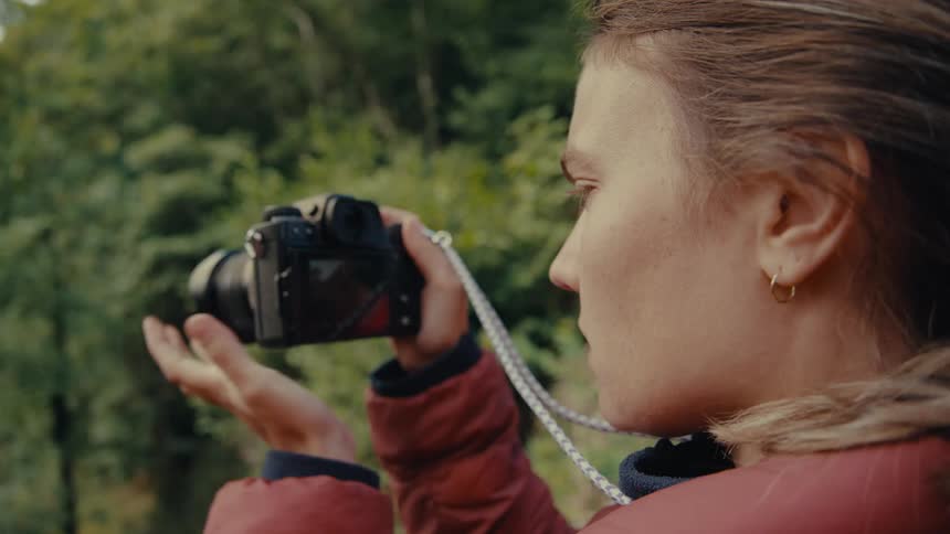 A person stands outdoors and holds a camera to look at the distance while another person stands behind them