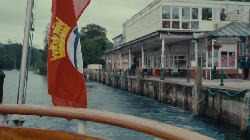 A boat with an orange flag featuring a yellow crown and a white circle departs a lakeside pier where people are walking along a raised wooden deck with white support columns