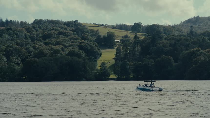 The video shows a wide shot of a small white and blue motorboat moving across a lake in the background a lush green landscape with a forest and a grassy hill can be seen