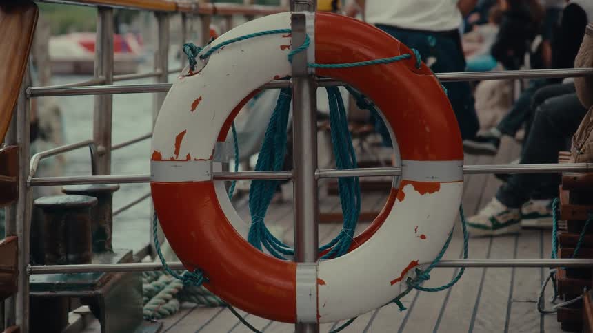A large white and orange life preserver hangs on the railing of a boat with a blue rope wrapped around it and blurred people seated in the background