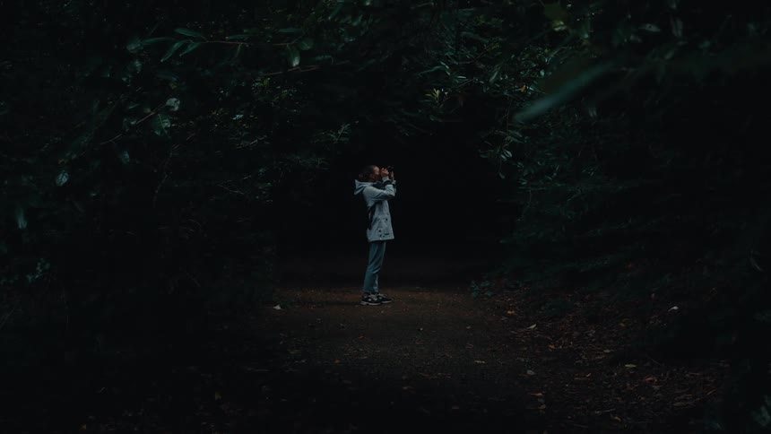 A person stands on a path in a forest and holds a device to their face while looking away from the camera