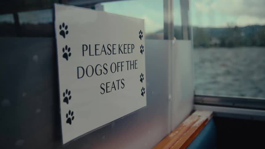 A sign that says please keep dogs off the seats is visible inside of a boat the boat travels on a body of water with a lush green mountain range visible through the window