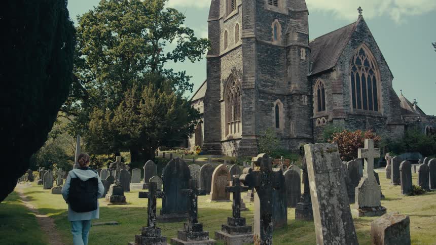 A person with light hair and a backpack is walking away from the camera on a dirt path through a graveyard with many dark and weathered gravestones all in front of a tall stone church