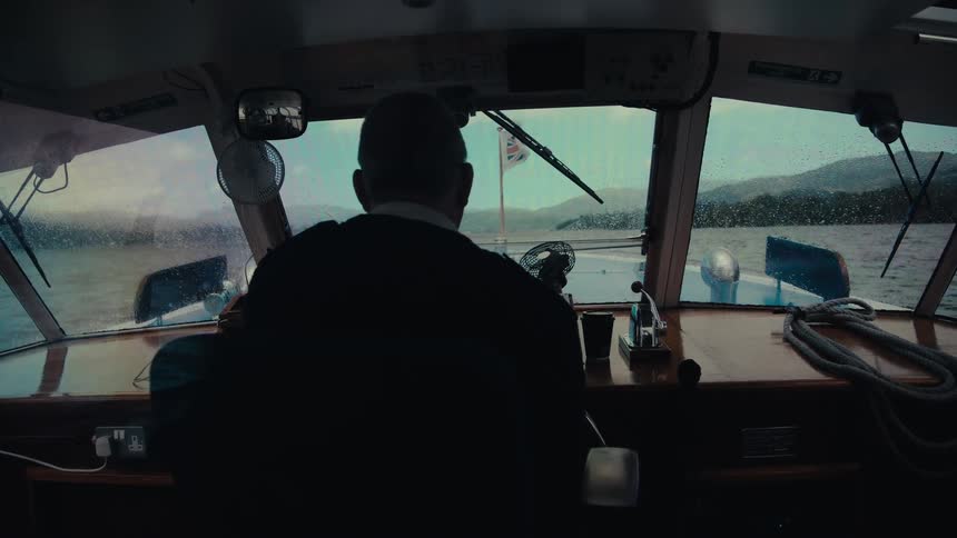 A man with his back to the camera steers a boat on a body of water toward distant hills under a cloudy sky with a union jack flag hanging from the front and a windshield wiper moving back and forth