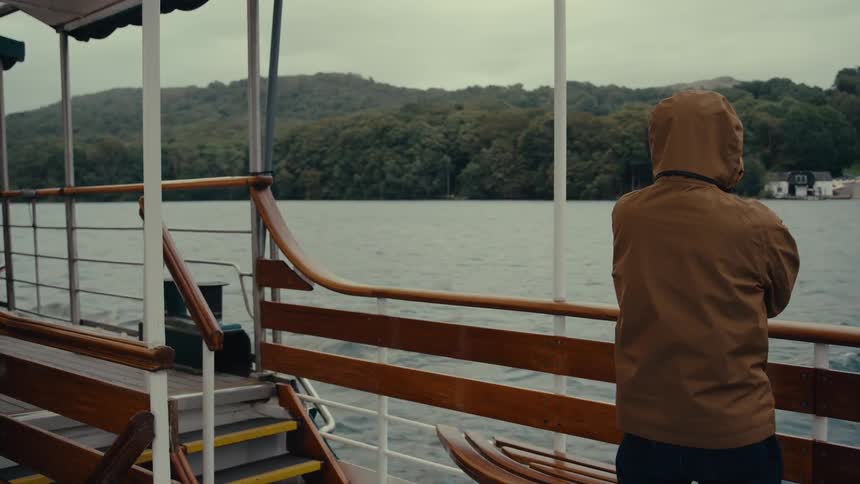 Person in a brown hooded jacket looks at the landscape while standing on a boat over the lake