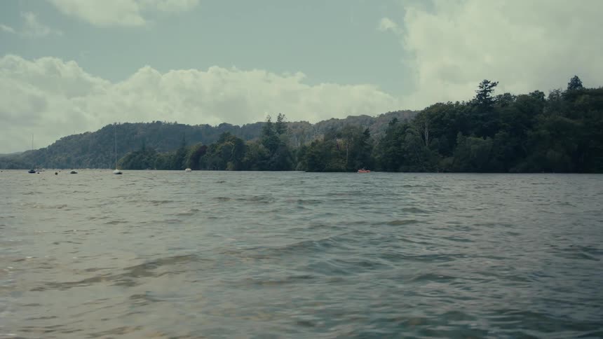 Lake water in the foreground with treecovered hills in the background under a partly cloudy sky