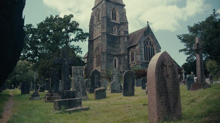 A graveyard with a variety of gravestones and monuments in a grassy field in front of a tall dark stone church with a steeple and large gothic windows