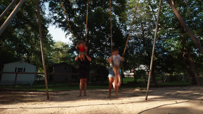 A family of four at a park with the mother and father pushing their two children on swings next to one another