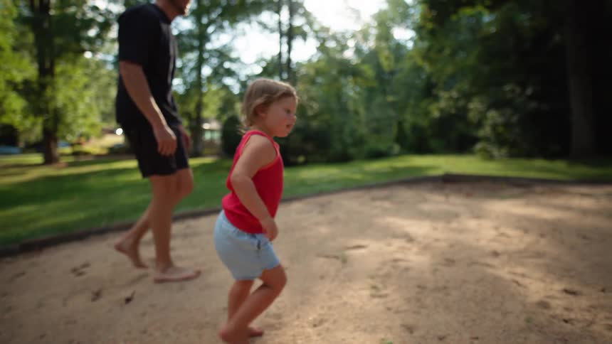 A man with a shaved head and tattoos on his arms is seen helping a small child who is wearing a red shirt and denim shorts walk on a sandy playground the man is holding the childs waist and encouraging them to take steps