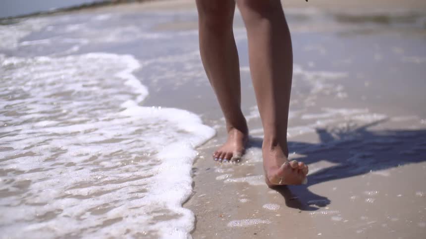 A tight shot of a young woman walking through wet sand and water as waves gently roll on shore