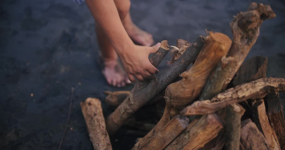 A person with bare feet stands on dark sand next to a pile of driftwood reaching down to add another piece to the stack