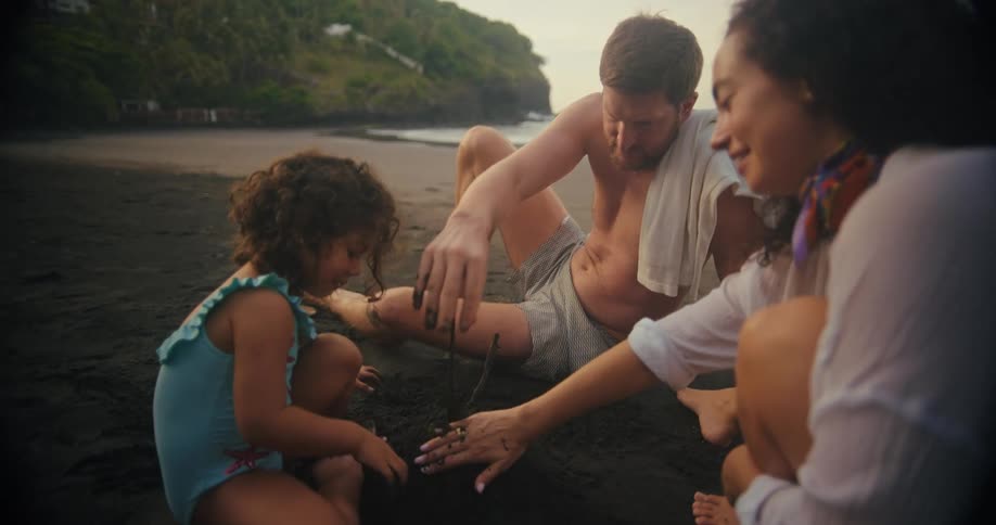 A family of three enjoys an evening on a dark sand beach focused on building a sandcastle together they sit closely with a young child playing in the sand while the parents watch and assist