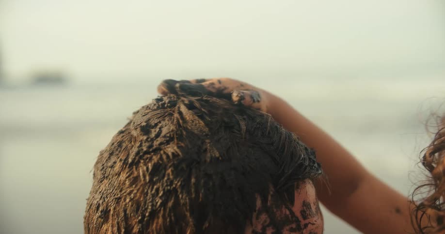 A person with a mud covered head is seen from behind as another persons muddy hands massage their hair