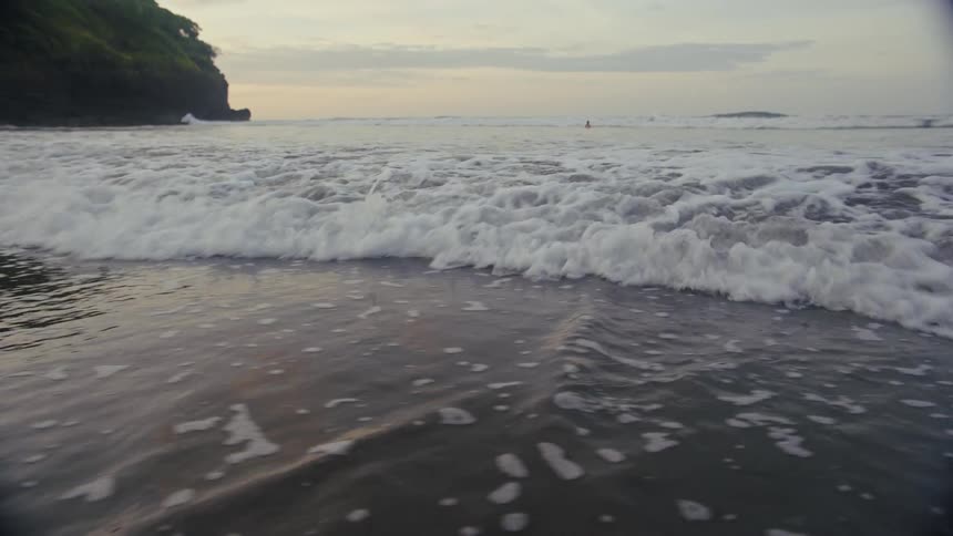 A lowangle shot captures the gentle waves of the ocean rolling onto a dark sand beach at dusk the camera is positioned just above the waterline showing the water bubble and foam as it washes in and out