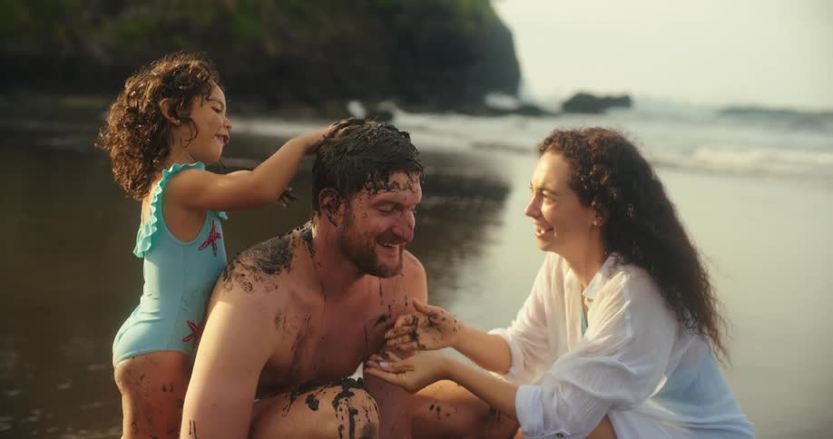 A smiling family is on a beach where a girl and a woman are covering a man in dark mud while he laughs