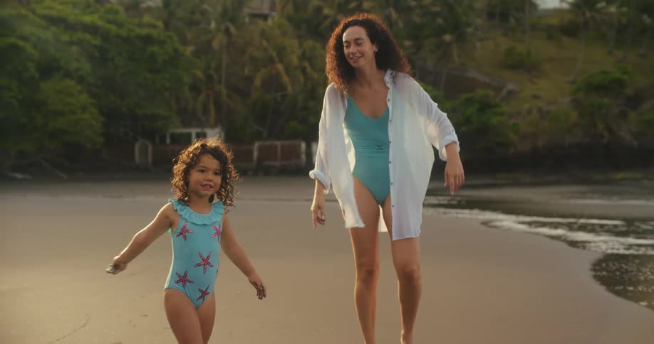 A mother and her young daughter playfully walk along the shore of a beach with dark sand the little girl points excitedly at something offcamera before turning and running toward the waves with her mother following close behind