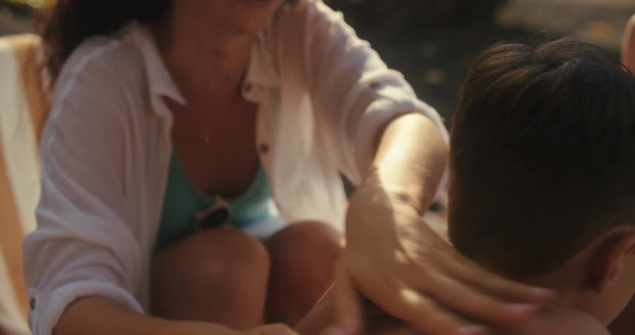 A woman in a light blue bikini and a white cover up is putting white sunscreen on a young boys back at the beach as she looks up at him and smiles