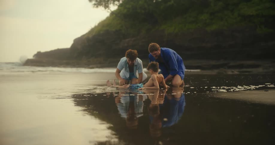 Two adults and a child play together at the seashore with their reflection in the shallow water