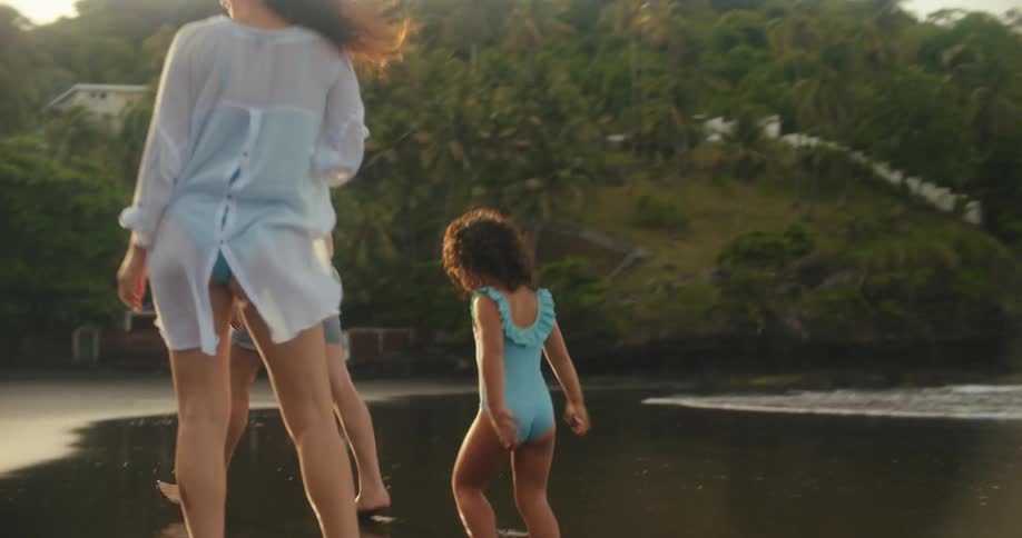The video shows a family of three a man a woman and a young girl standing on a beach with a lush green hillside in the background the man and the girl are smiling as they stand in the shallow water