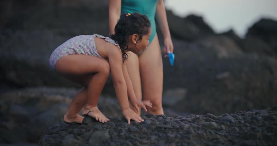 The video shows a young girl with brown hair in a swimsuit walking on a beach she is walking past some dark rocks and a blurry figure of a woman in a blue swimsuit is visible in the background