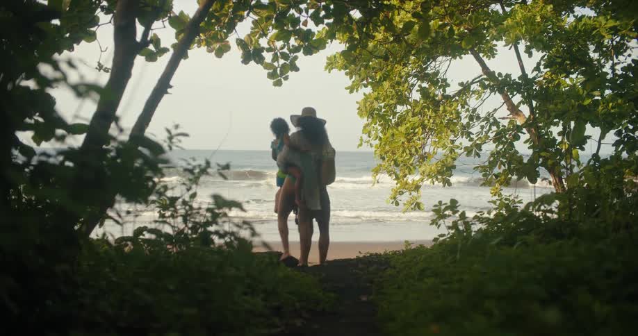 Mother carries her daughter while a family walks through a grove of trees in order to access the beach on a summer day