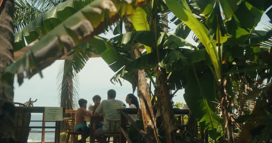 A family of four sits together at a wooden picnic table enjoying a meal or snack surrounded by lush tropical foliage the camera pans slowly from behind a curtain of broad green leaves revealing the group in a relaxed outdoor setting