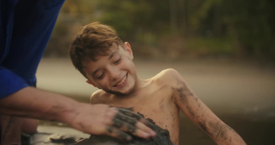 A person in a blue shirt uses their hands to cover a smiling boy with mud as he lies in a puddle