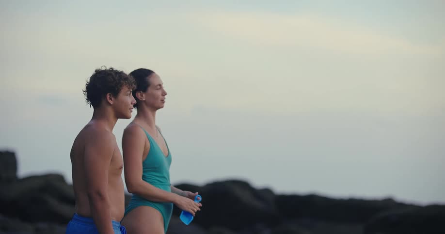 A young couple a man and a woman stands together on a rocky coastline at dusk looking out at the water they turn to each other with warm smiles before looking back out at the horizon