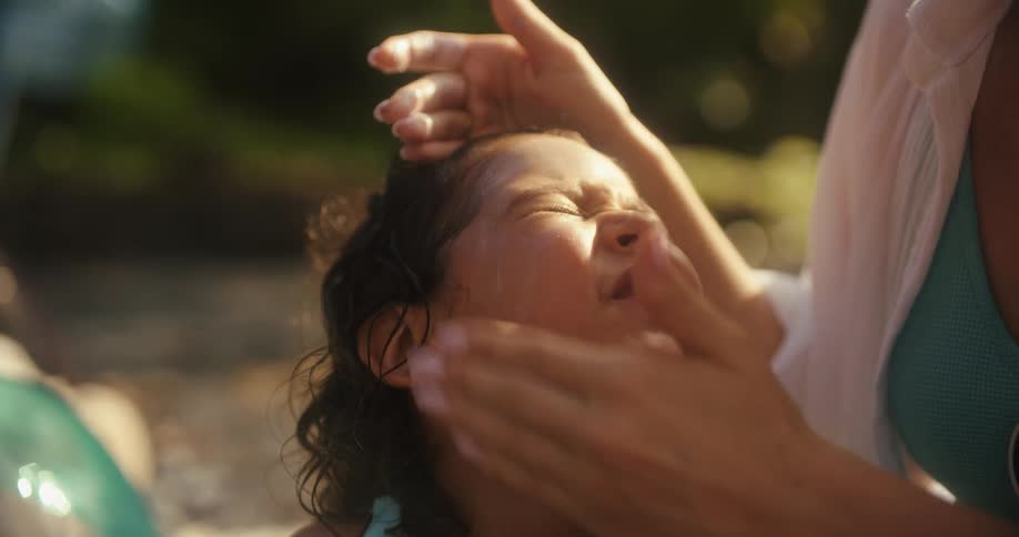 A mother gently applies sunscreen to her smiling childs face in the bright outdoor sun the child joyfully closes her eyes and laughs as the mother carefully rubs the lotion in