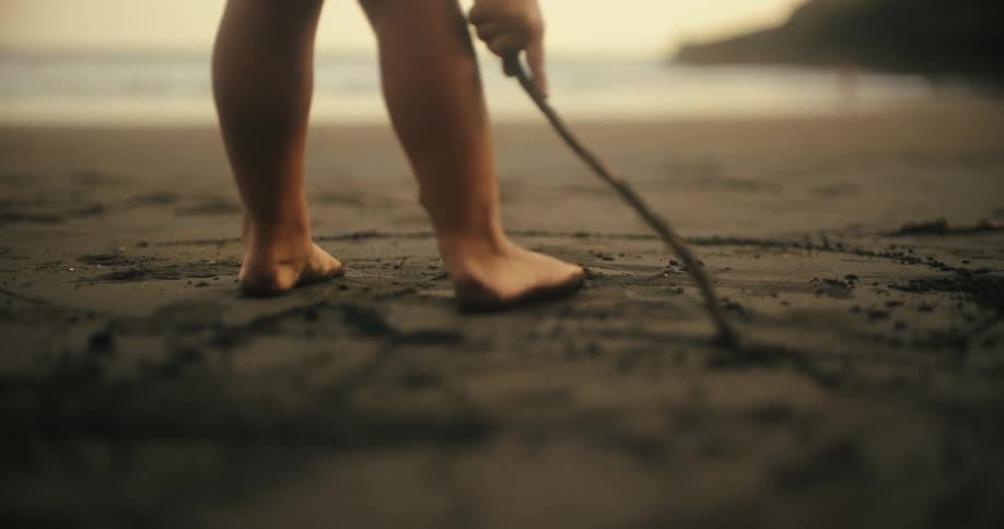A lowangle shot captures a persons bare feet walking across dark wet sand on a beach at dusk the person uses a stick to draw a line in the sand as they walk along the shoreline