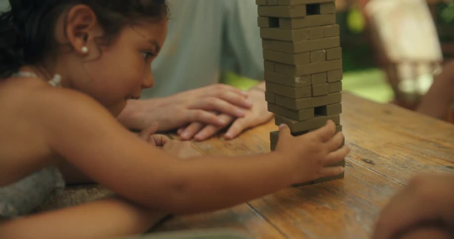 A young girl carefully pulls a block from the jenga tower her hand moving with intense focus her family members are gathered around her at a picnic table watching with anticipation