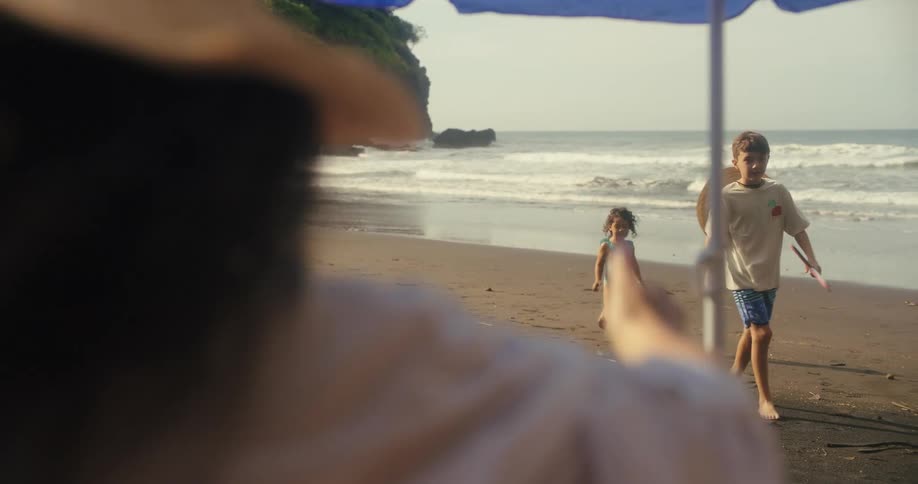 A shot from a low angle captures a family of three with two children and a man playing and walking on a dark sand beach they are enjoying their time on the shore with the ocean and a large green hillside in the background