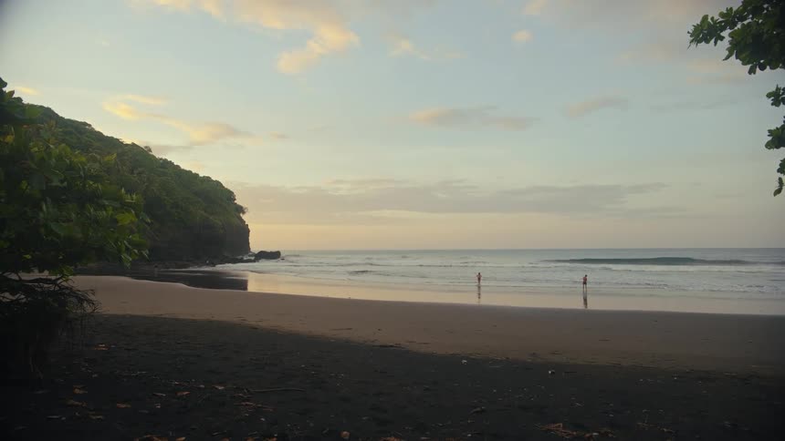 The camera captures a peaceful beach scene at dusk with gentle waves rolling onto the sand a lush treecovered cliff on the left and a few people in the distance complete the serene coastal view