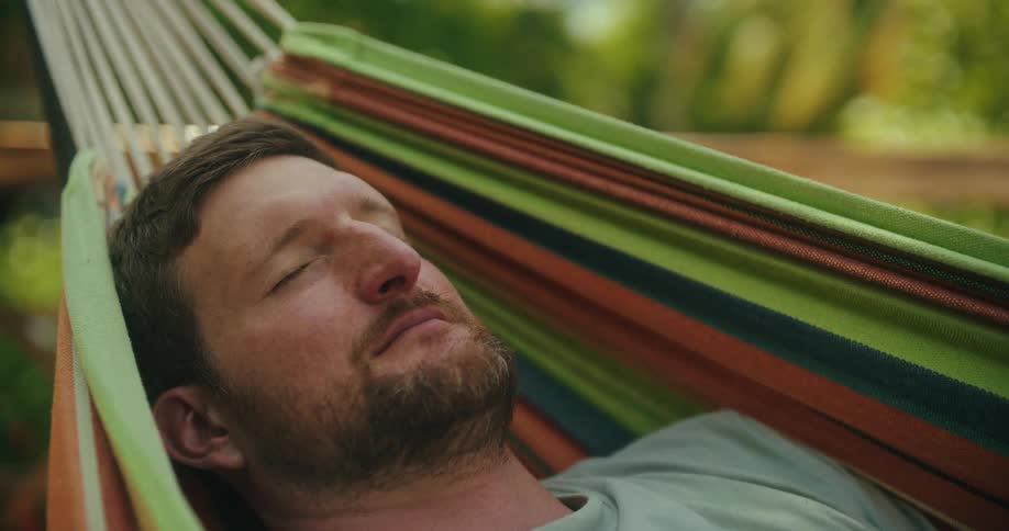 A man with a beard is sleeping peacefully in a colorful striped hammock he sways gently back and forth appearing completely relaxed and undisturbed