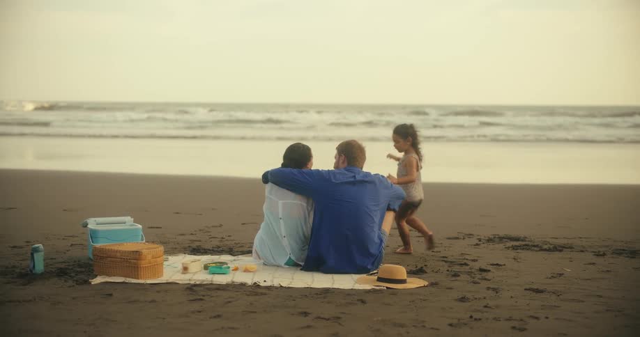 Father and mother kiss and cuddle during a beach picnic before their daughter runs to join them