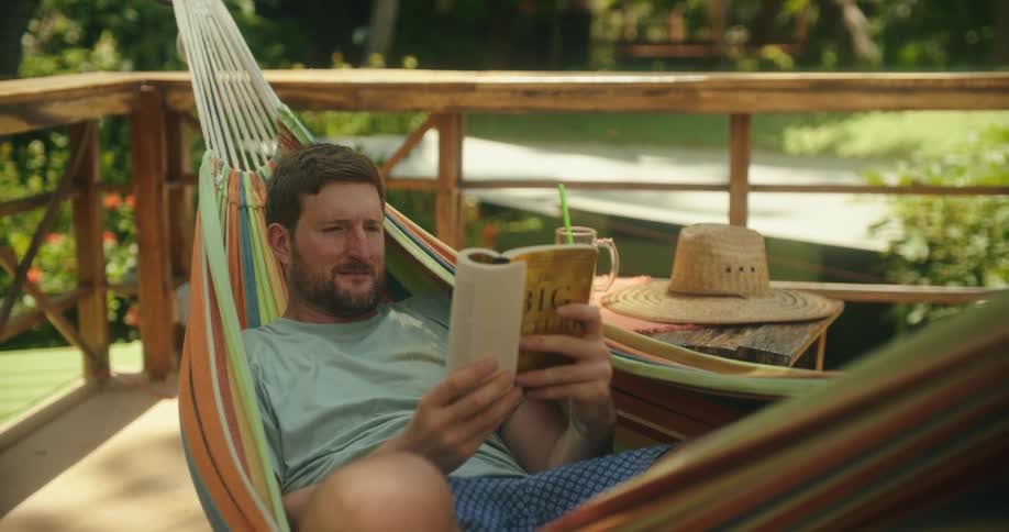 A man relaxes in a hammock reading a book with a drink nearby