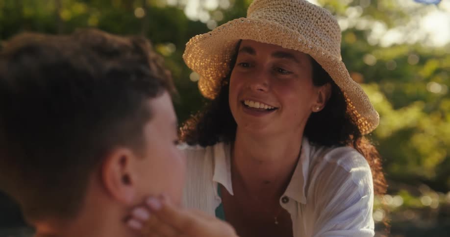 A smiling woman in a straw hat gently applies sunscreen to a young boys face while they are at the beach